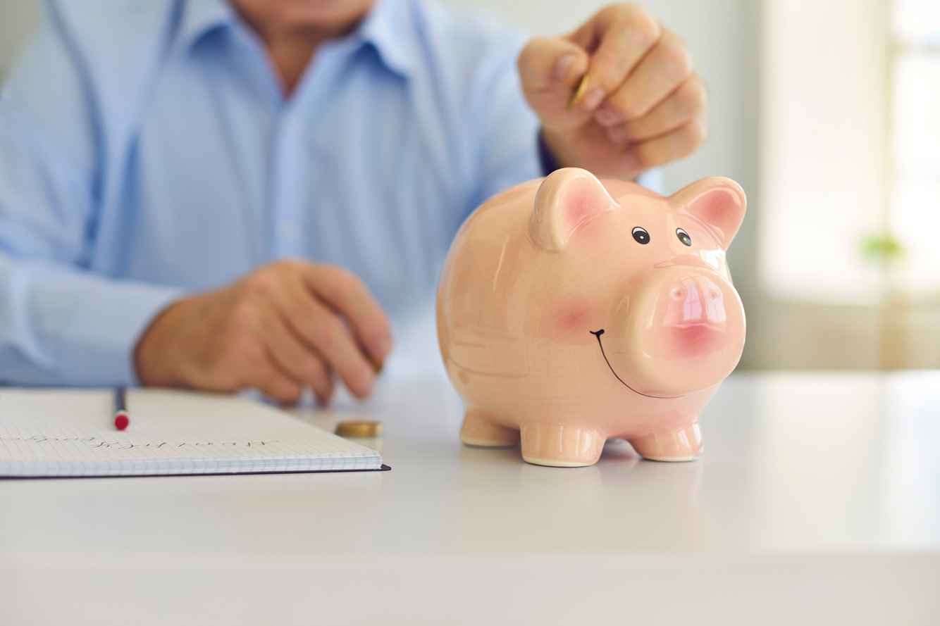 Senior Man Saving Money and Putting Coin in Cute Shiny Piggy Bank Standing on Desk